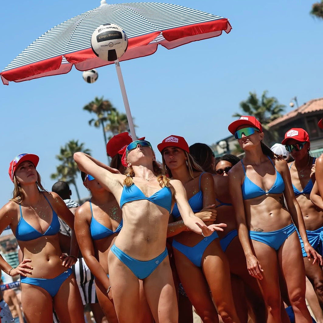 King of the Beach Miramar volleyball and Group of women in blue bikinis playing beach volleyball under a red and white umbrella.